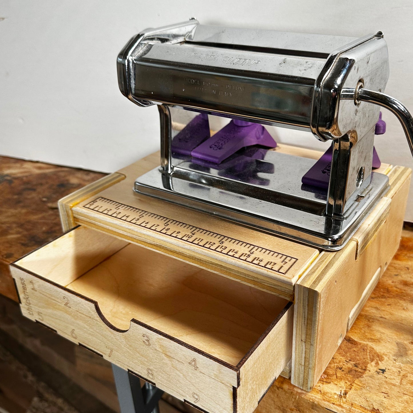 A front view of the pasta machine base with a pasta machine clamped in. The front drawer is open. The front panel of the drawer has spaces to record the thickness settings of the machine being used.